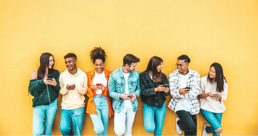A group of six diverse young adults lean against a bright yellow wall, engaged with their smartphones and enjoying each other's company.