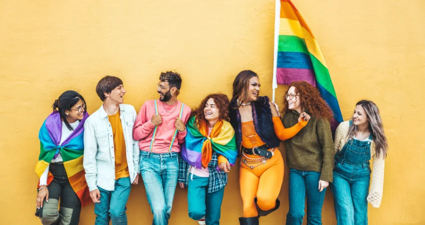 A group of people, draped in rainbow flags, celebrates diversity against a yellow wall.
